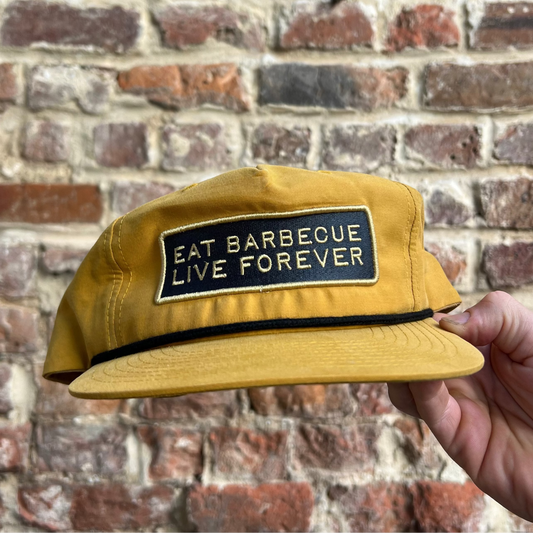 A yellow baseball cap with an embroidered patch that reads 'EAT BARBECUE LIVE FOREVER', held against a brick wall background.