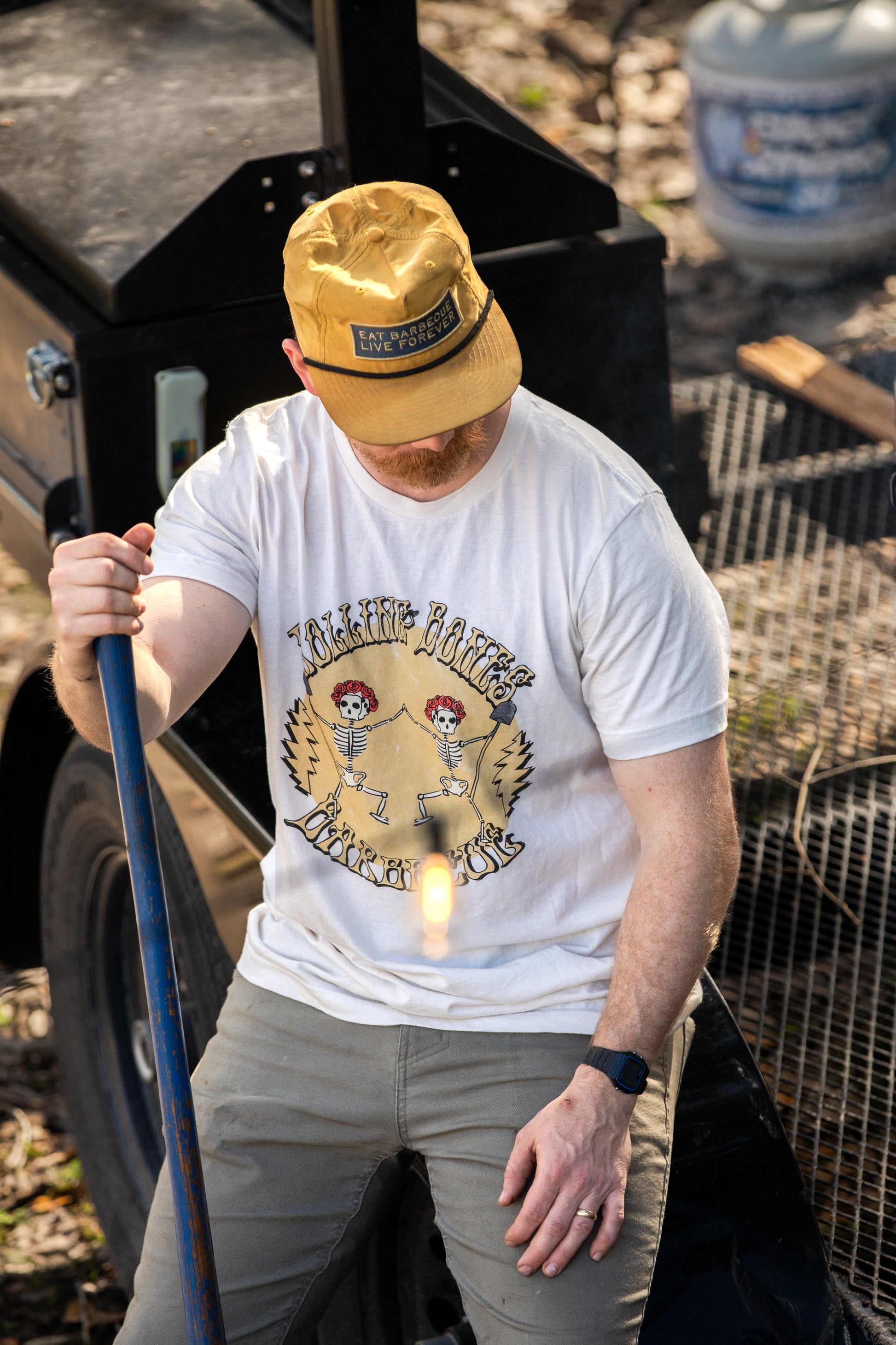 A person wearing a white crew neck T-shirt with a graphic print of a skeleton dancing and text, standing near outdoor equipment.