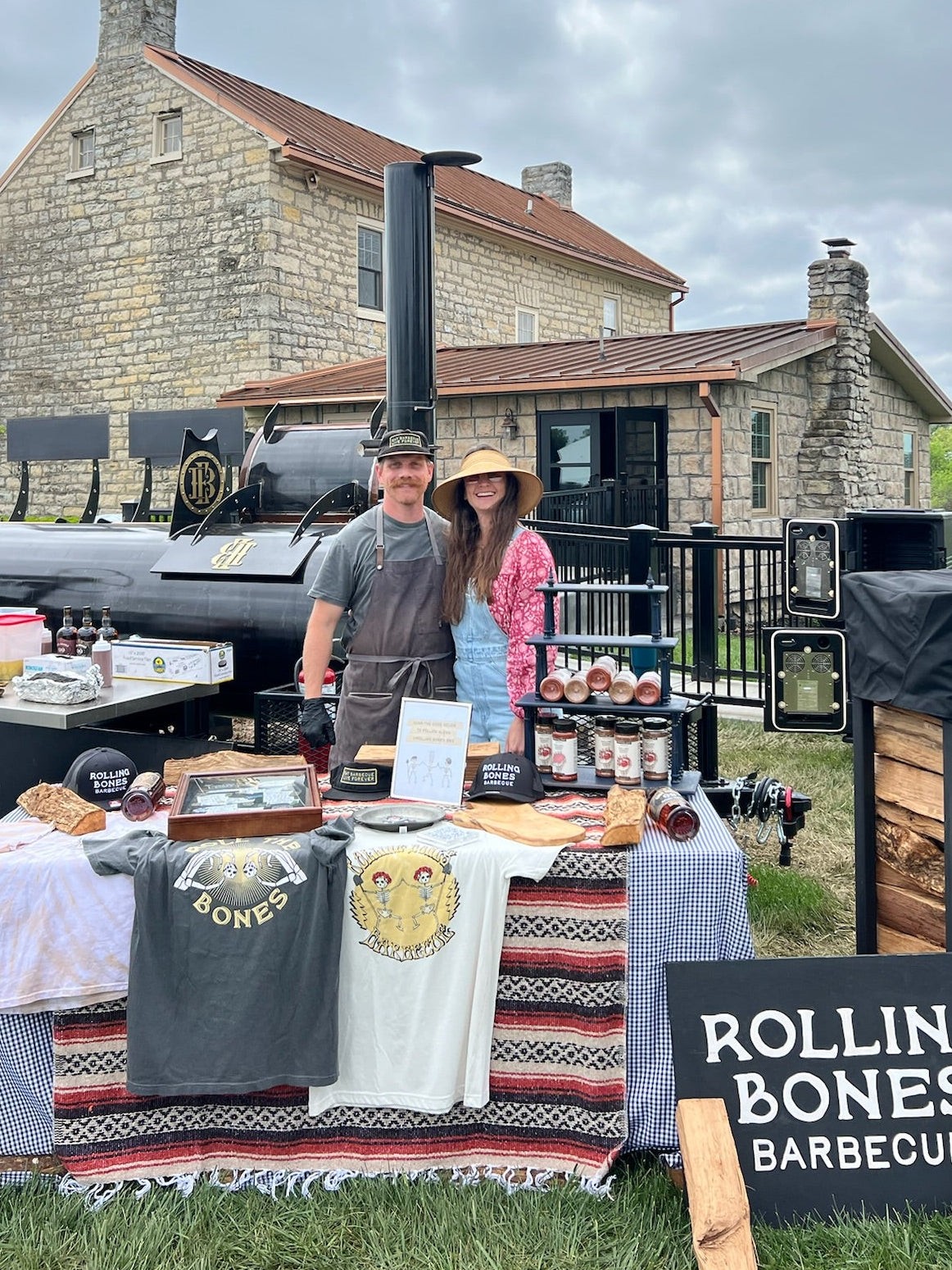 Will and Virginia standing behind their Rolling Bones barbecue setup with a sign reading 'Rolling Bones Barbecue' in front of a stone building at a cookout.
