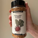Hand holding a jar of 'Rolling Bones Barbecue' Perpetual Veg dry rub seasoning  on a beige background