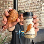 Person holding a roasted chicken leg and a container of 'Rolling Bones' seasoning against a brick wall.
