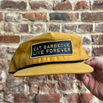 A yellow baseball cap with an embroidered patch that reads 'EAT BARBECUE LIVE FOREVER', held against a brick wall background.