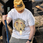 A person wearing a white crew neck T-shirt with a graphic print of a skeleton dancing and text, standing near outdoor equipment.