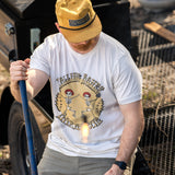 A person wearing a white crew neck T-shirt with a graphic print of a skeleton dancing and text, standing near outdoor equipment.