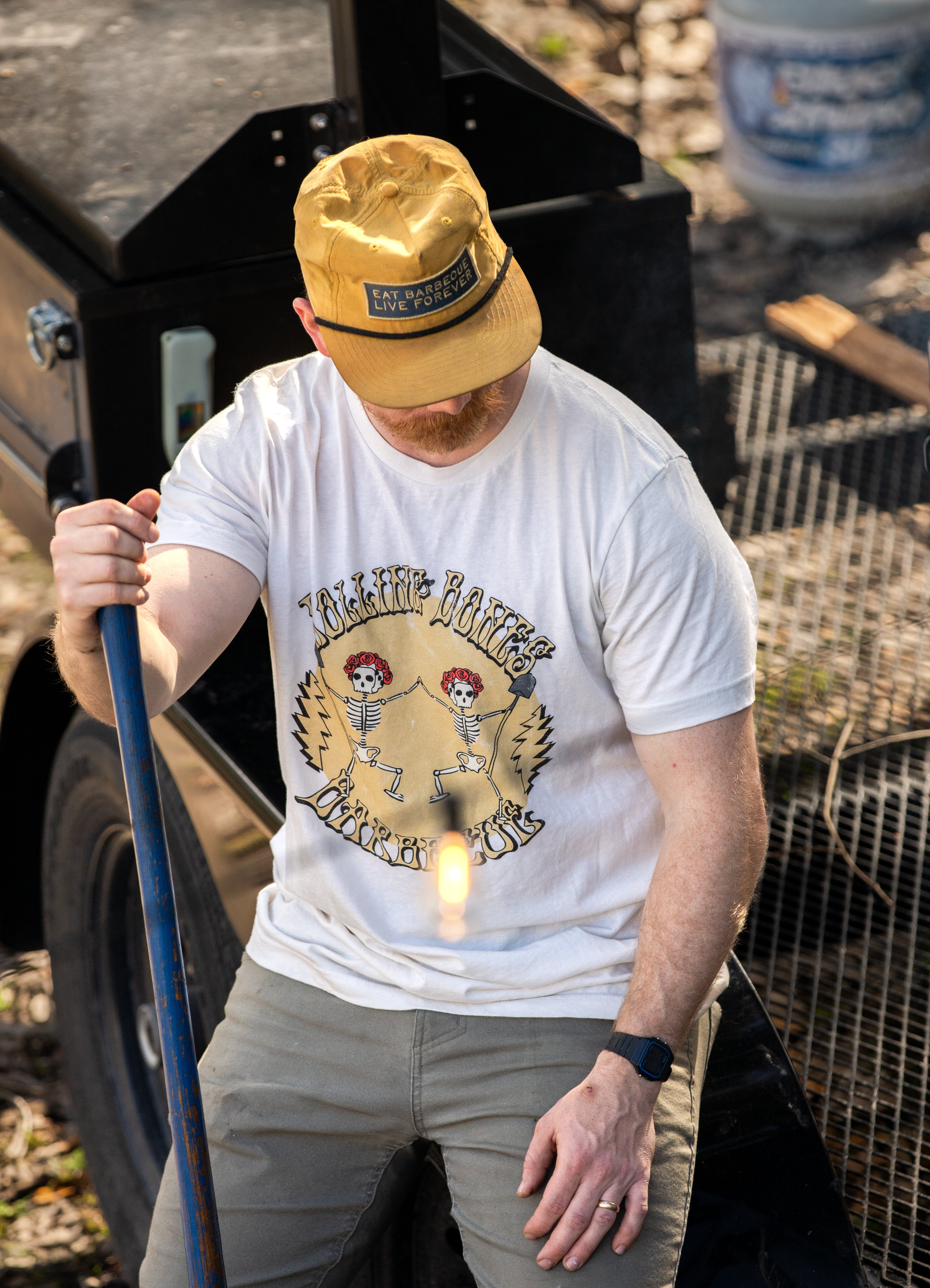 A person wearing a white crew neck T-shirt with a graphic print of a skeleton dancing and text, standing near outdoor equipment.