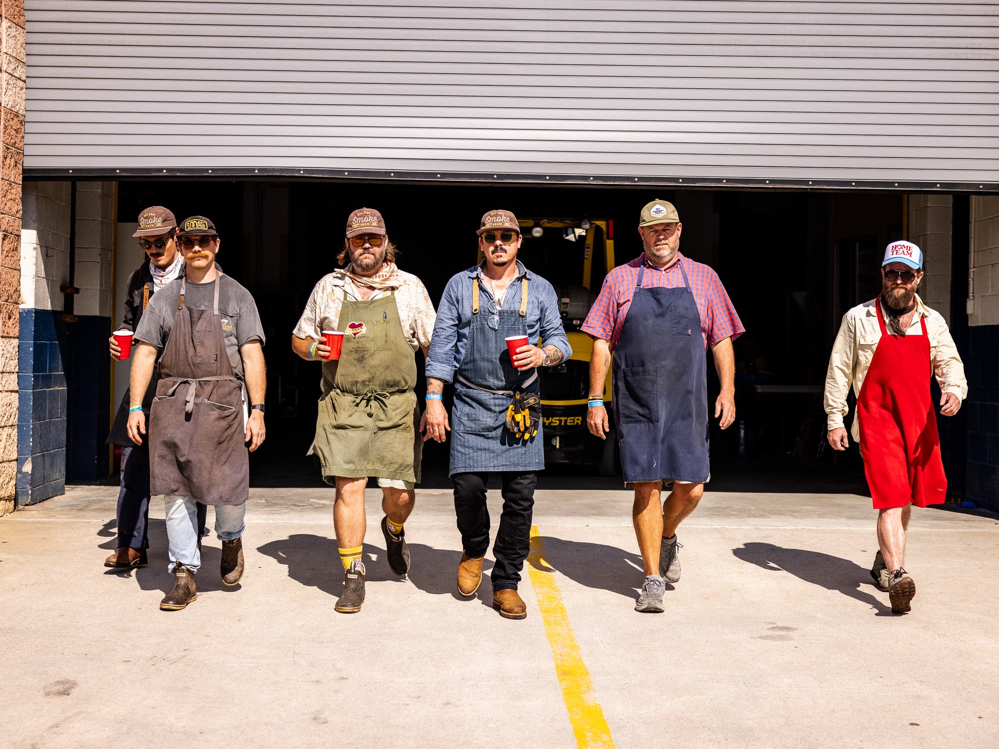A gathering of the Rolling Bones BBQ "Really Good" community at a live event. The group is comprised of six people wearing aprons and hats walking out of a garage getting ready to smoke meats and host a barbecue event together.
