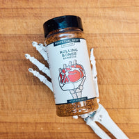 A bottle of of Rolling Bones Perpetual Beef dry rub seasoning sitting on a wooden surface with a skeleton hand holding it.