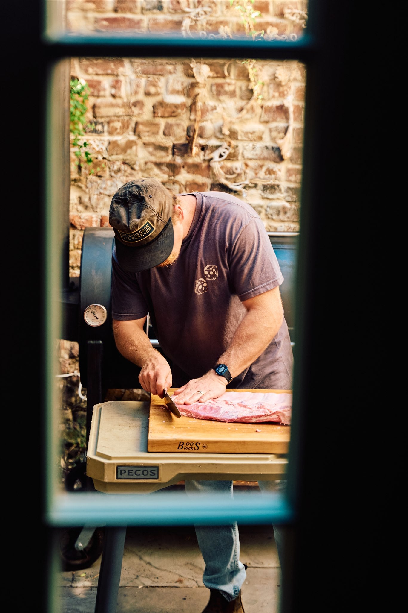 Will from Rolling Bones Barbecue trimming a rack of ribs in front of his barbecue smoker. Will is wearing a black Rolling Bones BBQ hat and a gray Double Die Rolling Bones BBQ t-shirt.