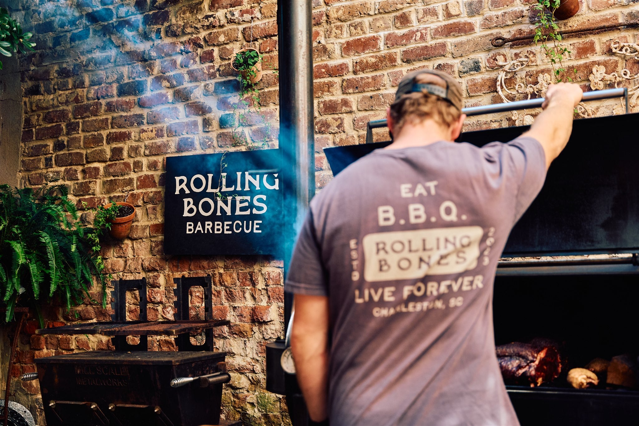Will from Rolling Bones BBQ working with a barbecue smoker at a Rolling Bones Barbecue community event, with a brick wall and sign in the background that says "ROLLING BONES BARBECUE"