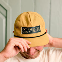 Person wearing a mustard-yellow Rolling Bones BBQ cap with 'Eat Barbecue Live Forever' text against a light green wall.