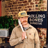 Will of Rolling Bones BBQ holding a skeleton hand prop in front of a brick wall with a  'Rolling Bones Barbecue' sign. He is wearing a mustard-yellow Rolling Bones BBQ hat that says "Eat Barbecue Live Forever"