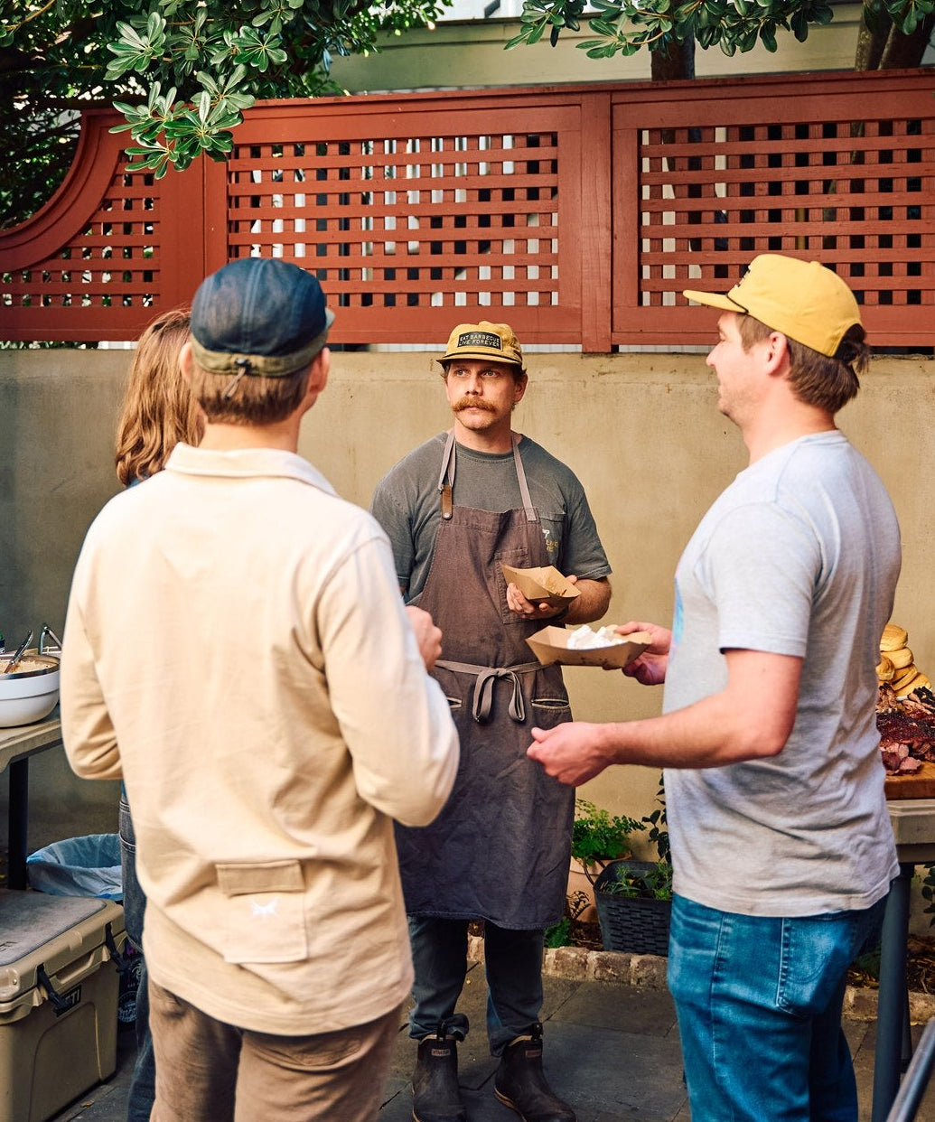 A gathering of the Rolling Bones BBQ "Really Good" community at a live event. The group of people is gathered around a grill and a barbecue smoker in an outdoor setting with trees and a wooden fence.