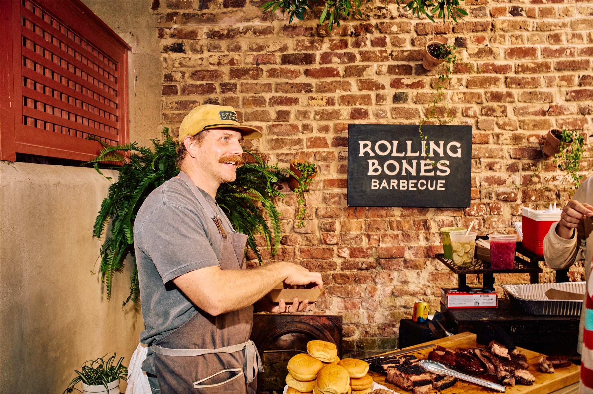 Will from Rolling Bones BBQ preparing food at a barbecue event for the Rolling Bones BBQ "Really Good" community. There is a black 'Rolling Bones Barbecue' sign on brick wall.