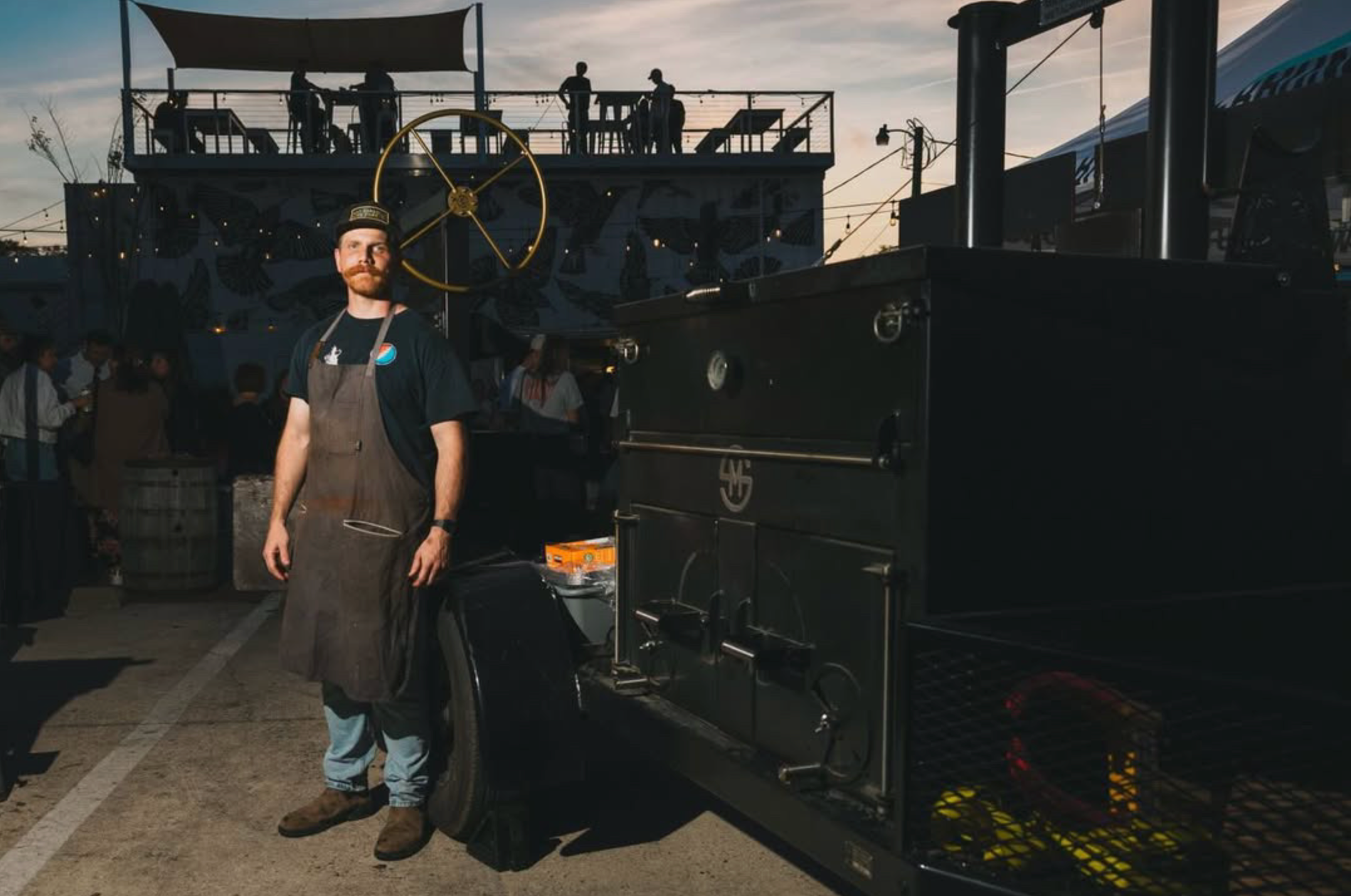 Will in an apron standing next to the smoker trailer rig at a barbecue event with a sunset in the background