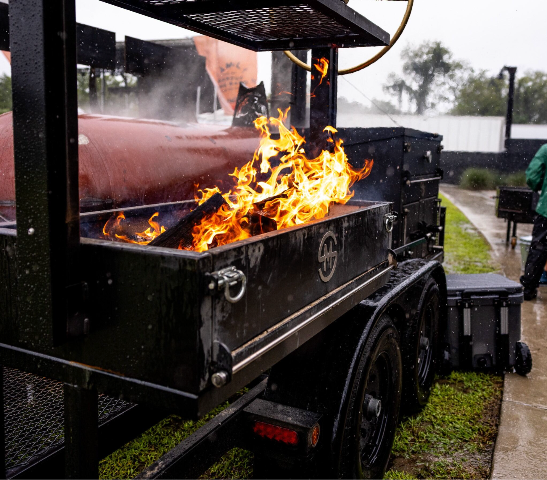 A barbecue grill with flames inside, outdoors on a rainy day. The barbecue smoker is from Mill Scale Metalworks.