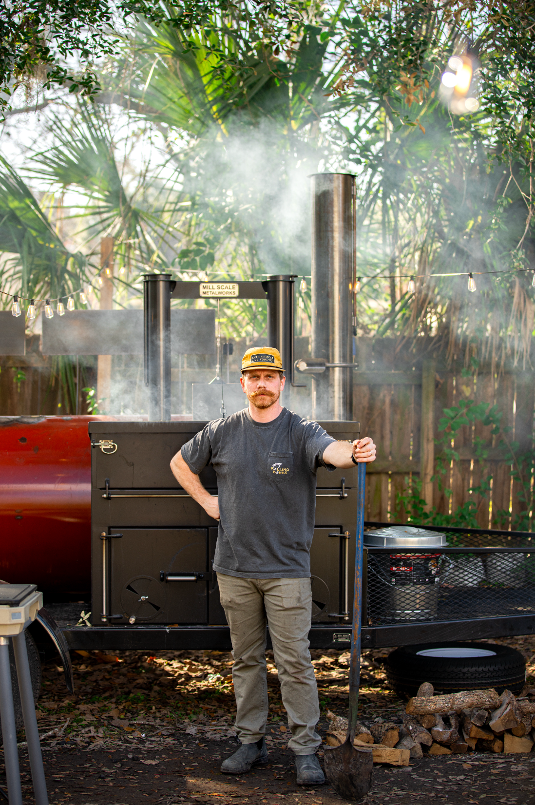 Will standing next to a Mill Scale barbecue smoker with smoke billowing in a garden setting